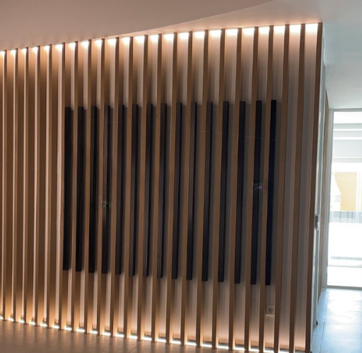 Revestimiento de pared moderno en recepción de oficina con lamas de madera y un panel central de perfilería metálica. Letras corpóreas de acero inoxidable retroiluminadas.